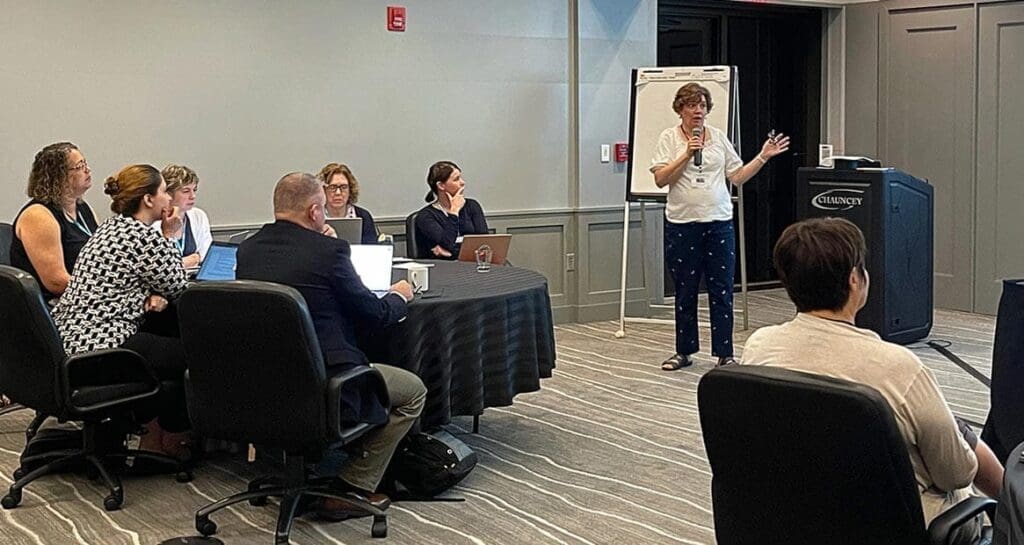 Kathleen Hill presenting in front of a podium to seated people at roundtables in front of her. The audience members are facing her and her presentation.